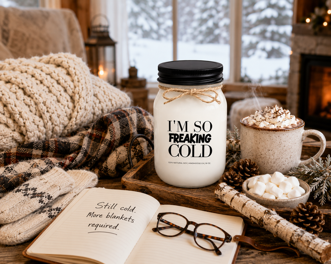 I’M SO FREAKING COLD soy candle styled beside a winter journal, reading glasses, hot chocolate, wool scarf, mittens, pinecones, and frosted greenery. Bathed in soft winter light, the scene reflects the cozy comforts of the season and the humorous reality of always feeling cold during winter.