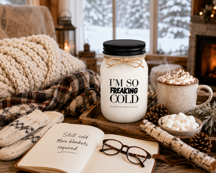 I’M SO FREAKING COLD soy candle styled beside a winter journal, reading glasses, hot chocolate, wool scarf, mittens, pinecones, and frosted greenery. Bathed in soft winter light, the scene reflects the cozy comforts of the season and the humorous reality of always feeling cold during winter.