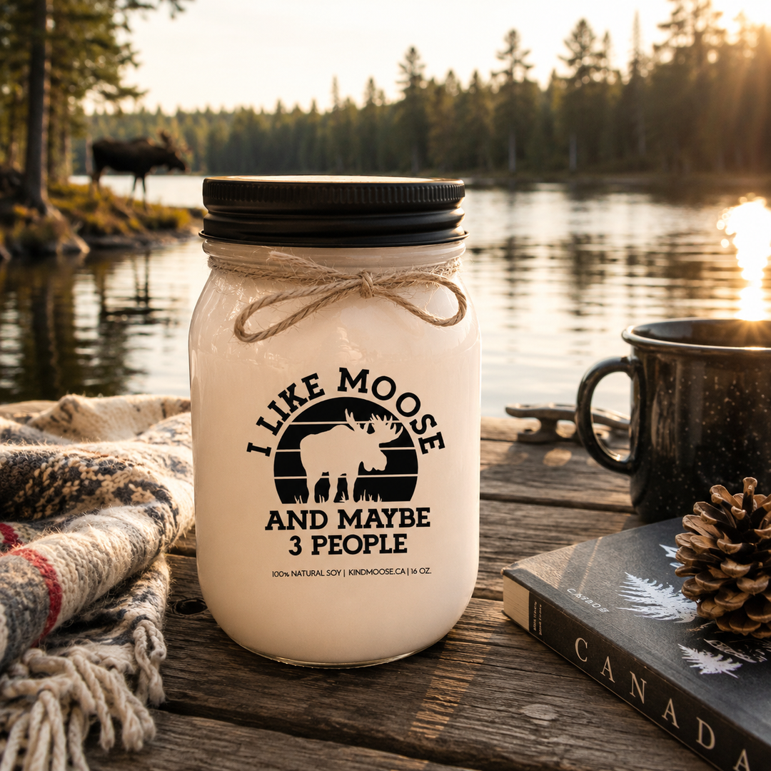 I Like Moose And Maybe 3 People candle in a frosted white mason jar with black lid displayed on a rustic cottage dock overlooking a peaceful Canadian lake surrounded by pine trees at sunrise.