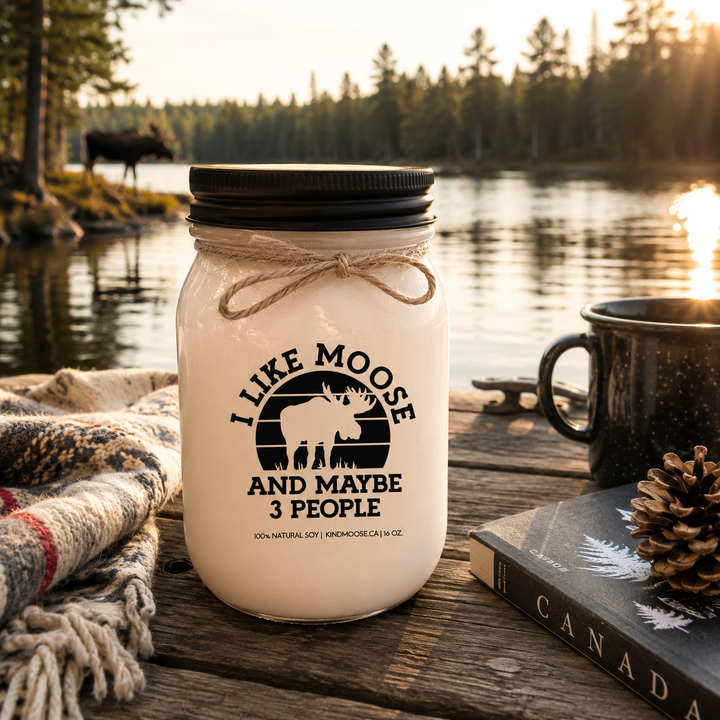 I Like Moose And Maybe 3 People candle in a frosted white mason jar with black lid displayed on a rustic cottage dock overlooking a peaceful Canadian lake surrounded by pine trees at sunrise.