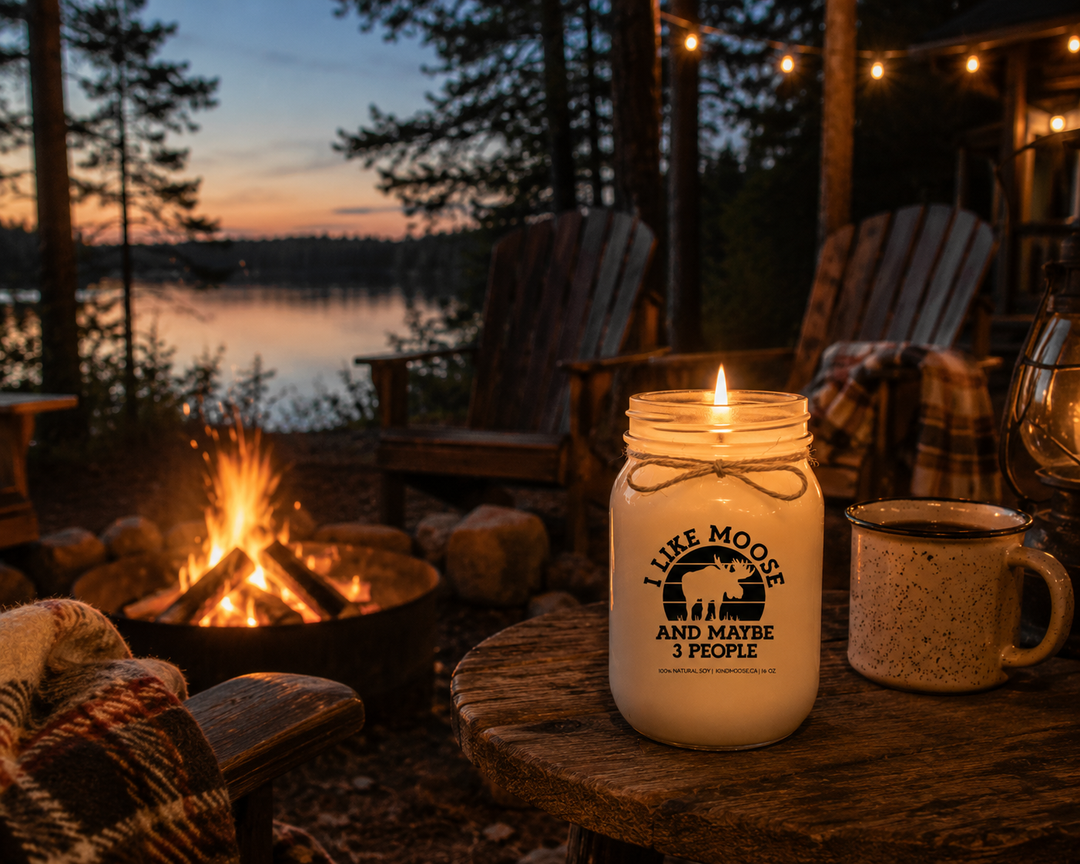 Burning I Like Moose And Maybe 3 People candle in a frosted white mason jar beside a campfire at a lakeside cottage with Adirondack chairs, string lights, and pine trees at dusk.