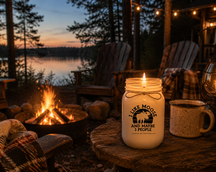 Burning I Like Moose And Maybe 3 People candle in a frosted white mason jar beside a campfire at a lakeside cottage with Adirondack chairs, string lights, and pine trees at dusk.