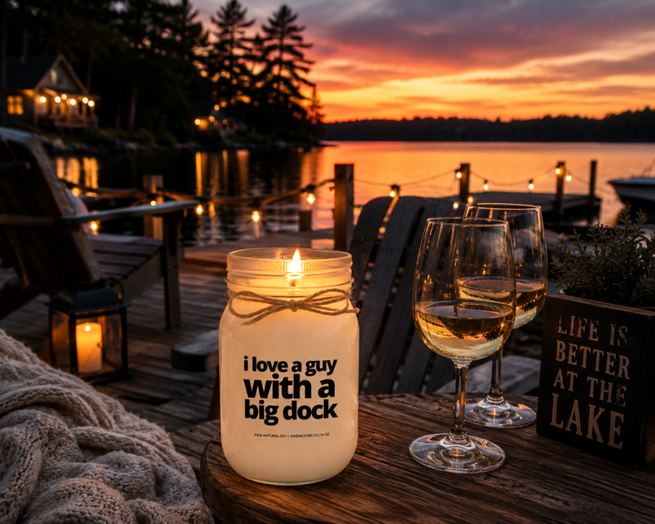 Burning I Love A Guy With A Big Dock soy candle on a lakeside cottage deck at sunset with Muskoka chairs, string lights, and peaceful lake views.