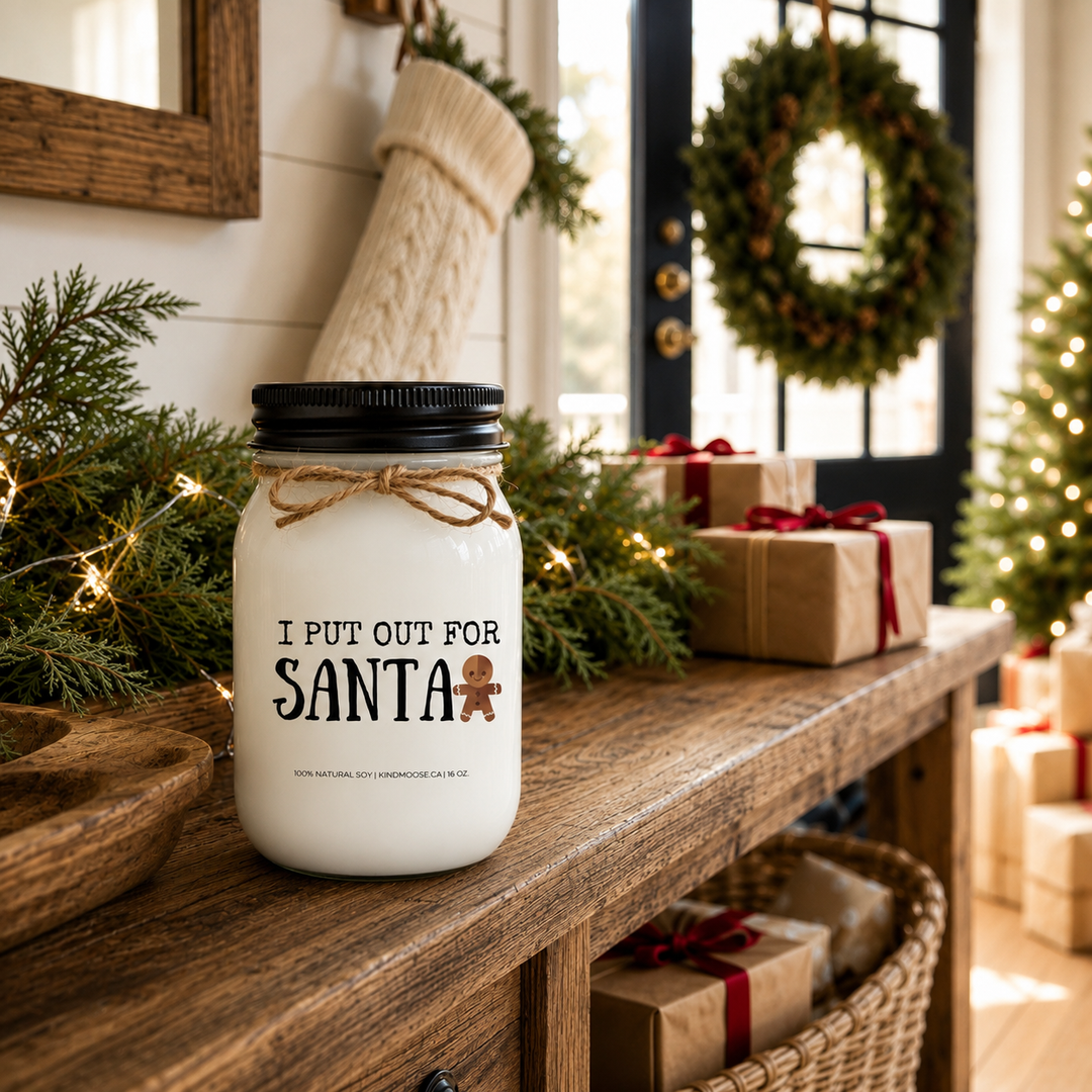 KINDMOOSE "I Put Out for Santa" 16 oz frosted white soy candle with a black lid displayed on a rustic farmhouse entry table decorated with cedar garland, wrapped Christmas gifts, a knit stocking and a holiday wreath, creating a warm and festive Christmas welcome.
