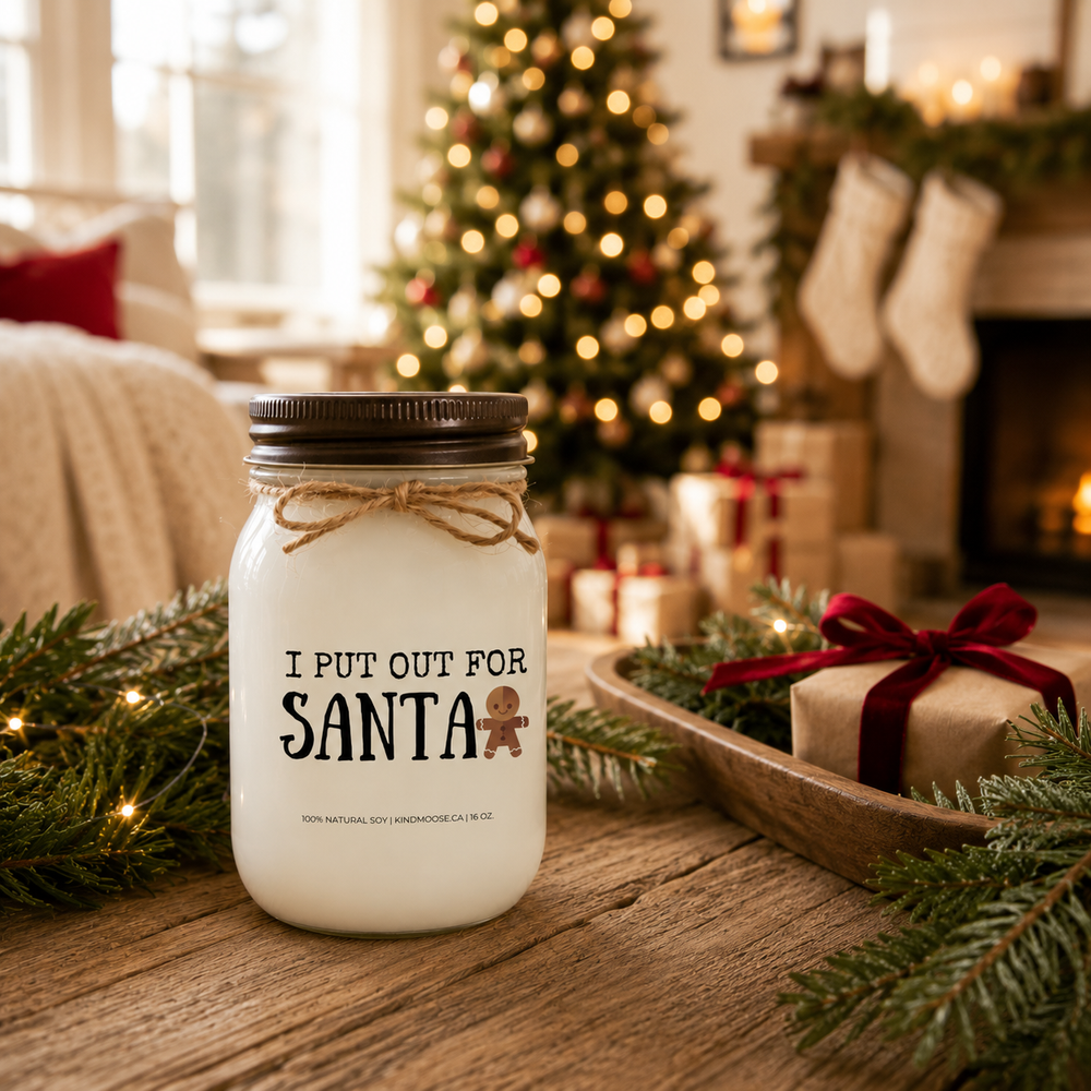 KINDMOOSE "I Put Out for Santa" 16 oz frosted white soy candle with a distressed bronze lid styled in a festive Christmas living room beside a beautifully wrapped holiday gift, cedar greenery and glowing Christmas tree lights.