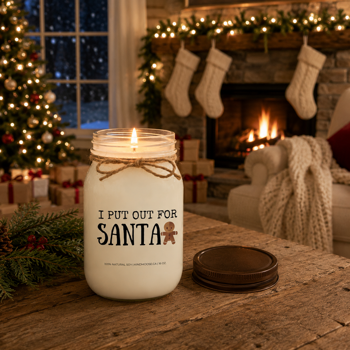 KINDMOOSE "I Put Out for Santa" burning 16 oz frosted white soy candle with the distressed bronze lid beside it on a rustic coffee table in front of a glowing fireplace and Christmas tree on Christmas Eve.