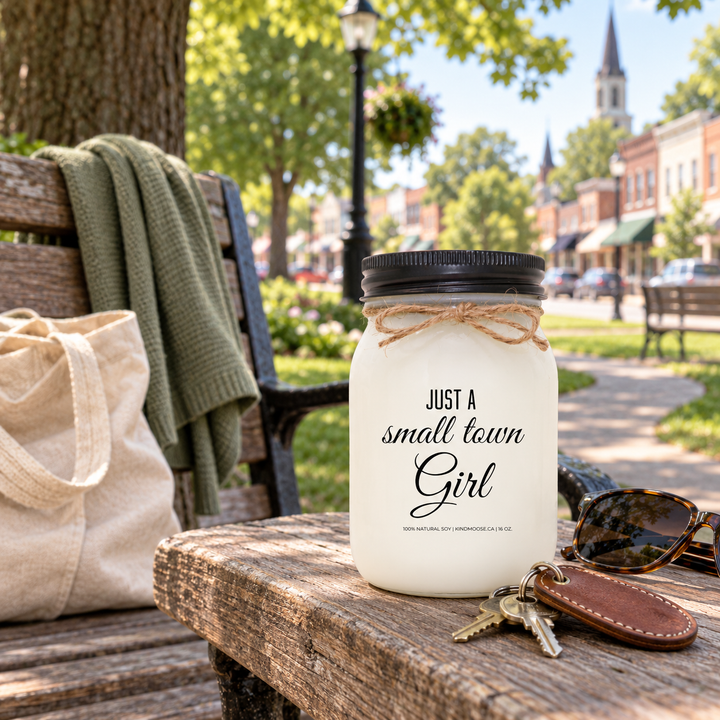 Just a Small Town Girl KINDMOOSE 16 oz candle with black lid on a park bench overlooking a familiar Canadian small-town main street.