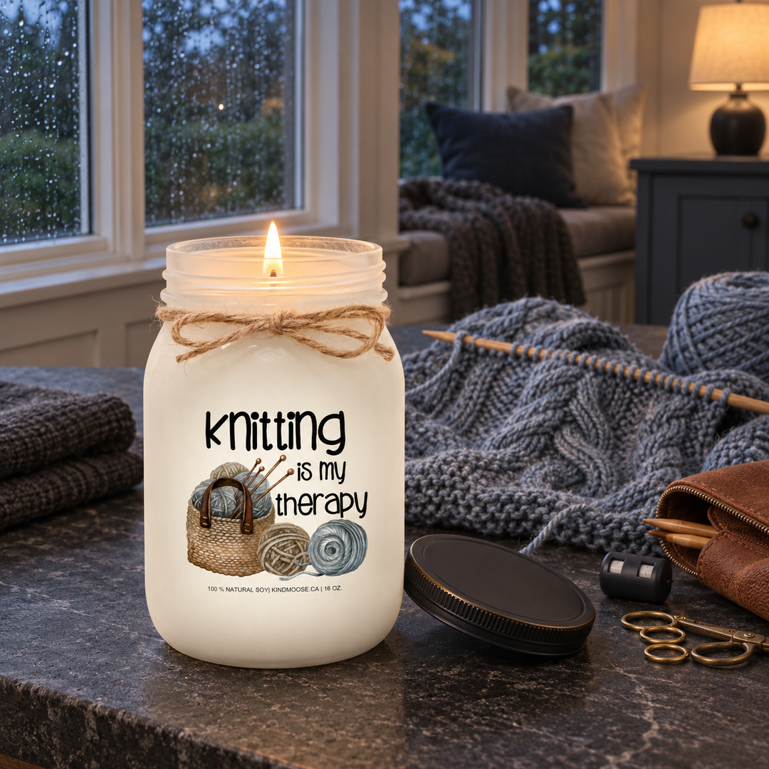 Burning KINDMOOSE Knitting Is My Therapy candle beside a blue-grey knitting project during a peaceful rainy evening.