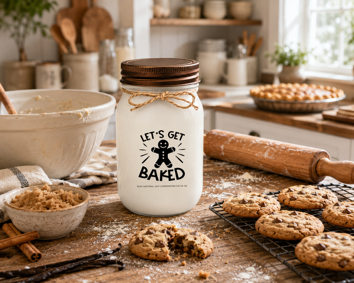 LET'S GET BAKED soy candle with distressed bronze lid styled on a farmhouse kitchen island beside freshly baked cookies, a cooling rack, mixing bowl, rolling pin, brown sugar, vanilla beans, and cinnamon sticks. The welcoming baking-day scene captures the joy of homemade treats and kitchen traditions shared with family and friends.