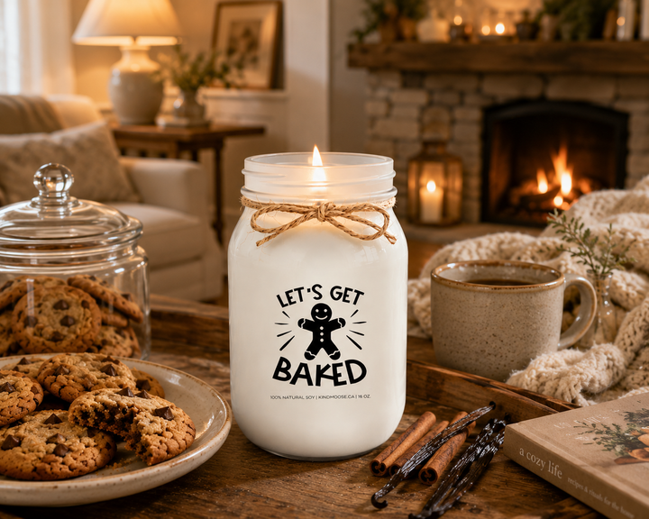 Burning LET'S GET BAKED soy candle displayed in a cozy farmhouse living room featuring a stone fireplace, fresh cookies, a cookie jar, knit blanket, rustic coffee table, and warm evening lighting. The inviting scene celebrates enjoying homemade treats, relaxing after a day of baking, and creating comforting family memories.