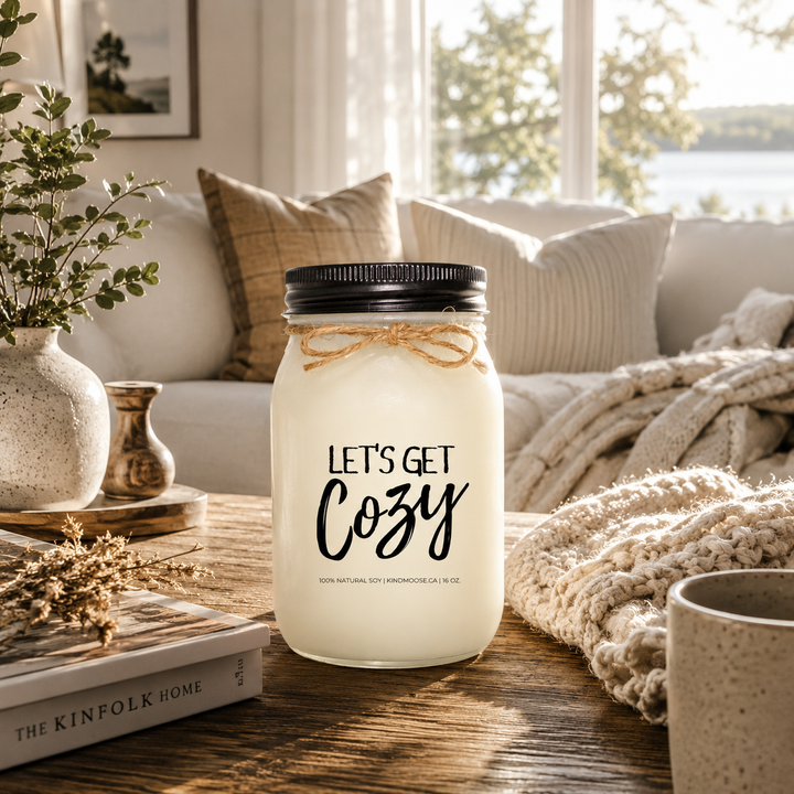 LET'S GET COZY inspirational soy candle by KINDMOOSE Candle Co. displayed on a rustic wood coffee table in a bright cottage living room with soft blankets, neutral pillows, and warm morning sunlight creating a cozy and relaxing atmosphere.