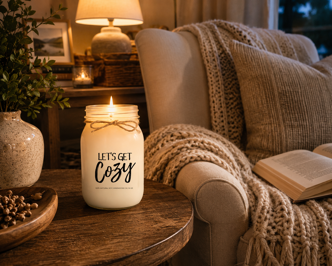 LET'S GET COZY inspirational soy candle by KINDMOOSE Candle Co. burning softly on a rustic wood side table beside a comfortable reading chair, knit blanket, open book, warm lamp glow, and cozy cottage décor in a peaceful evening setting.
