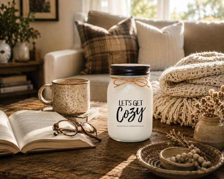 LET'S GET COZY inspirational soy candle by KINDMOOSE Candle Co. displayed on a rustic cottage coffee table beside an open novel, reading glasses, coffee mug, folded blanket, and warm natural sunlight creating a quiet and comforting moment at home.