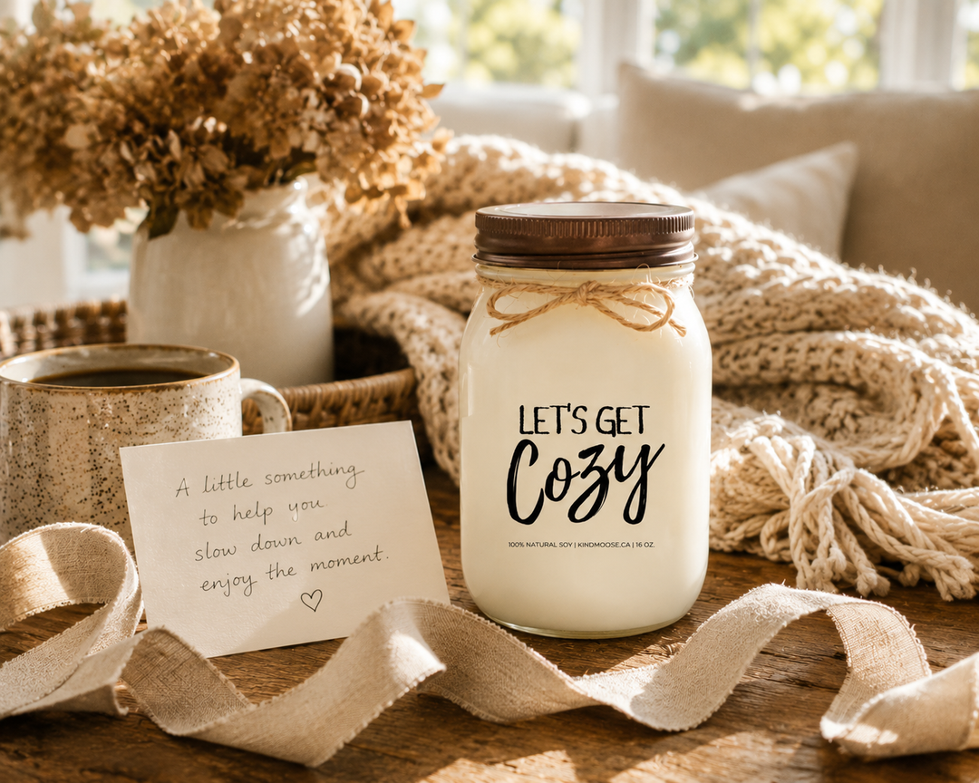 LET'S GET COZY inspirational soy candle by KINDMOOSE Candle Co. displayed as a thoughtful gift on a rustic wood table with a handwritten card, linen ribbon, cozy blanket, coffee mug, and warm afternoon sunlight creating a comforting housewarming gift scene.