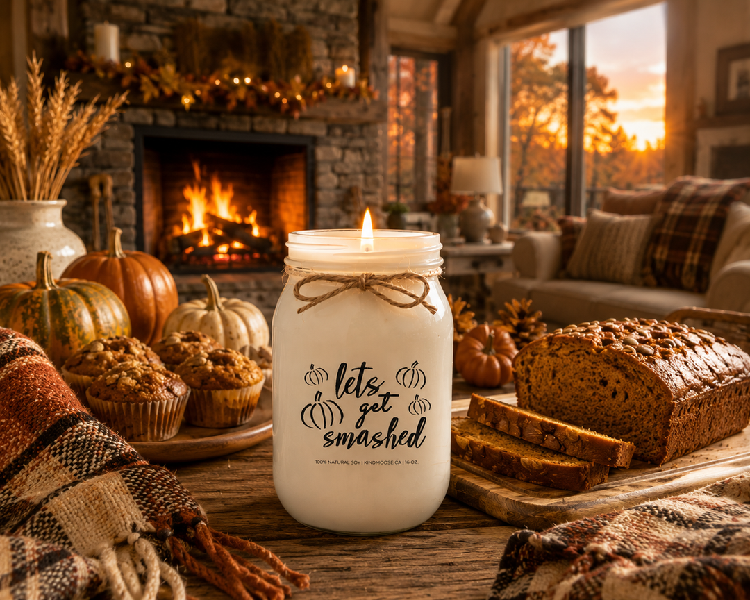 Burning LET'S GET SMASHED soy candle displayed in a cozy farmhouse living room featuring a stone fireplace, pumpkin loaf, pumpkin muffins, heirloom pumpkins, plaid blanket, pinecones, and warm evening lighting. The inviting autumn scene celebrates pumpkin season, comfort, and relaxing after a day of fall activities.