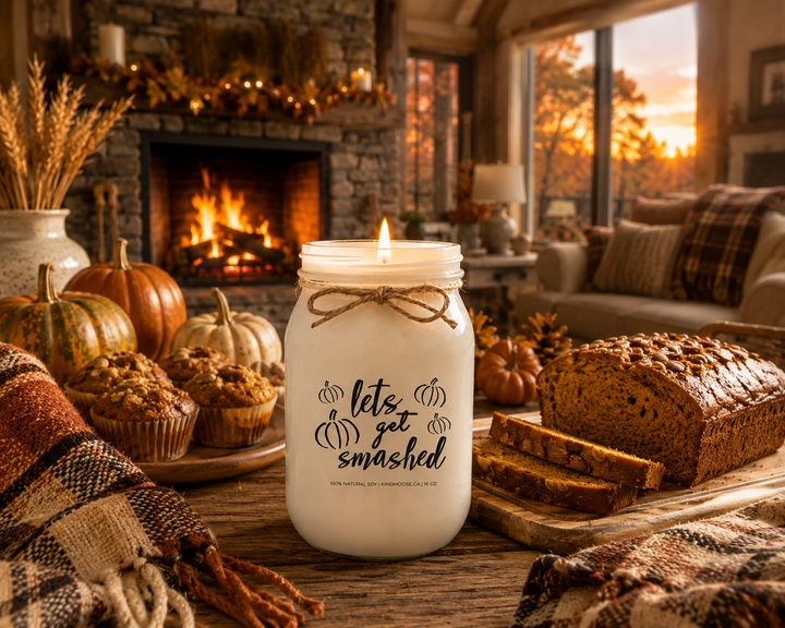 Burning LET'S GET SMASHED soy candle displayed in a cozy farmhouse living room featuring a stone fireplace, pumpkin loaf, pumpkin muffins, heirloom pumpkins, plaid blanket, pinecones, and warm evening lighting. The inviting autumn scene celebrates pumpkin season, comfort, and relaxing after a day of fall activities.