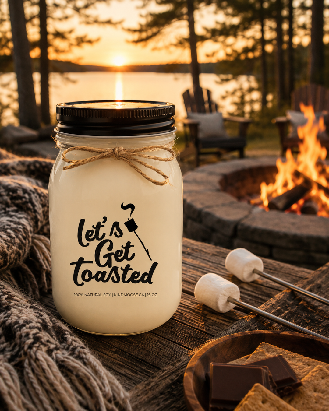 LET'S GET TOASTED soy candle by KINDMOOSE Candle Co. with black lid displayed on a rustic wood table beside an outdoor campfire at a lakeside cottage surrounded by pine trees during a warm sunset.