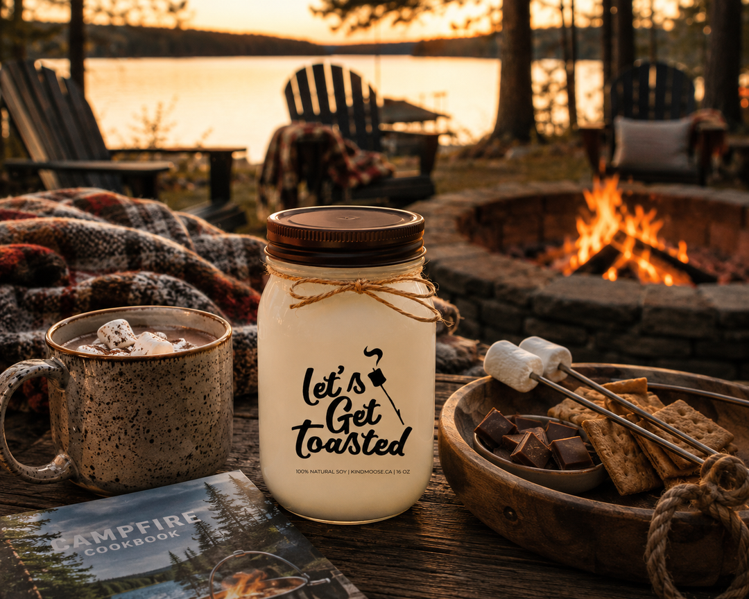 LET'S GET TOASTED soy candle with distressed bronze lid styled in a cozy cottage campfire gathering beside hot chocolate, a blanket, marshmallow roasting sticks, and a campfire cookbook overlooking the lake.