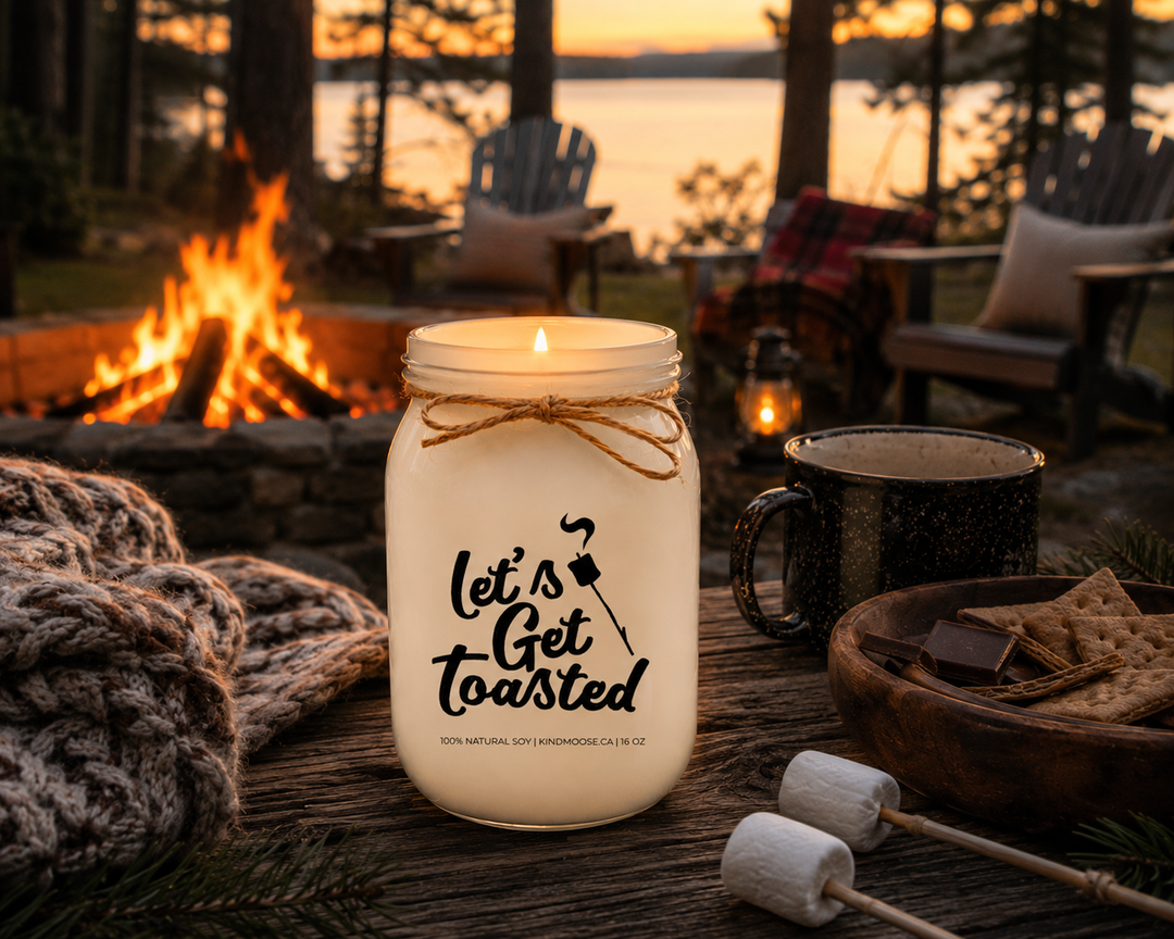 Burning LET'S GET TOASTED soy candle by KINDMOOSE Candle Co. on a rustic outdoor table beside a glowing campfire with Adirondack chairs, cozy blankets, pine trees, and warm evening light at a lakeside cottage.
