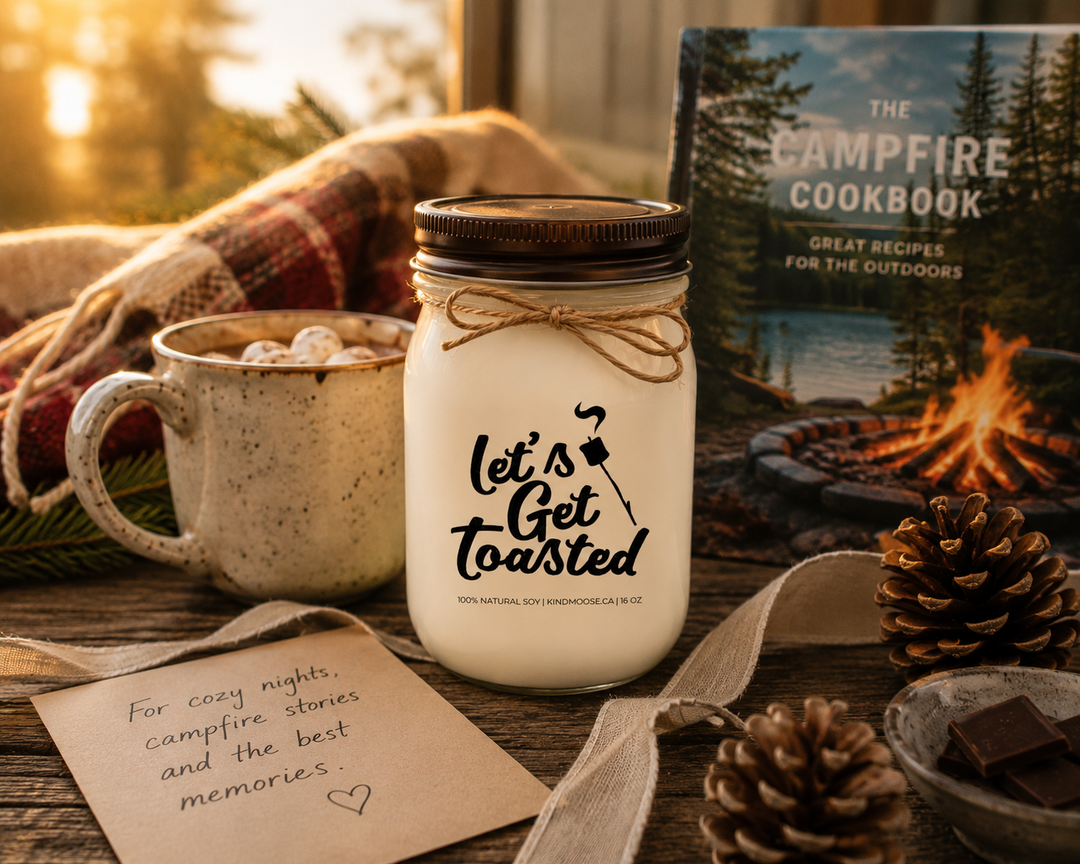 LET'S GET TOASTED soy candle with distressed bronze lid presented as a thoughtful cottage gift beside a handwritten card, linen ribbon, campfire cookbook, hot chocolate mug, pinecones, and warm golden sunlight.