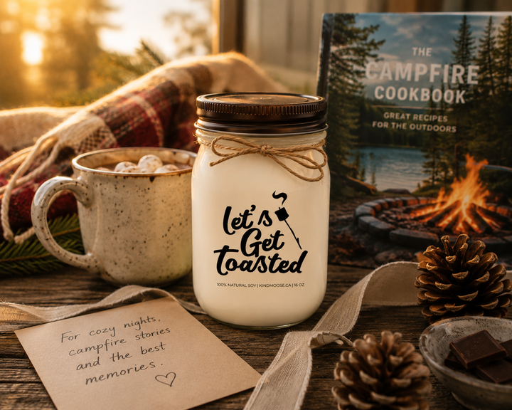 LET'S GET TOASTED soy candle with distressed bronze lid presented as a thoughtful cottage gift beside a handwritten card, linen ribbon, campfire cookbook, hot chocolate mug, pinecones, and warm golden sunlight.