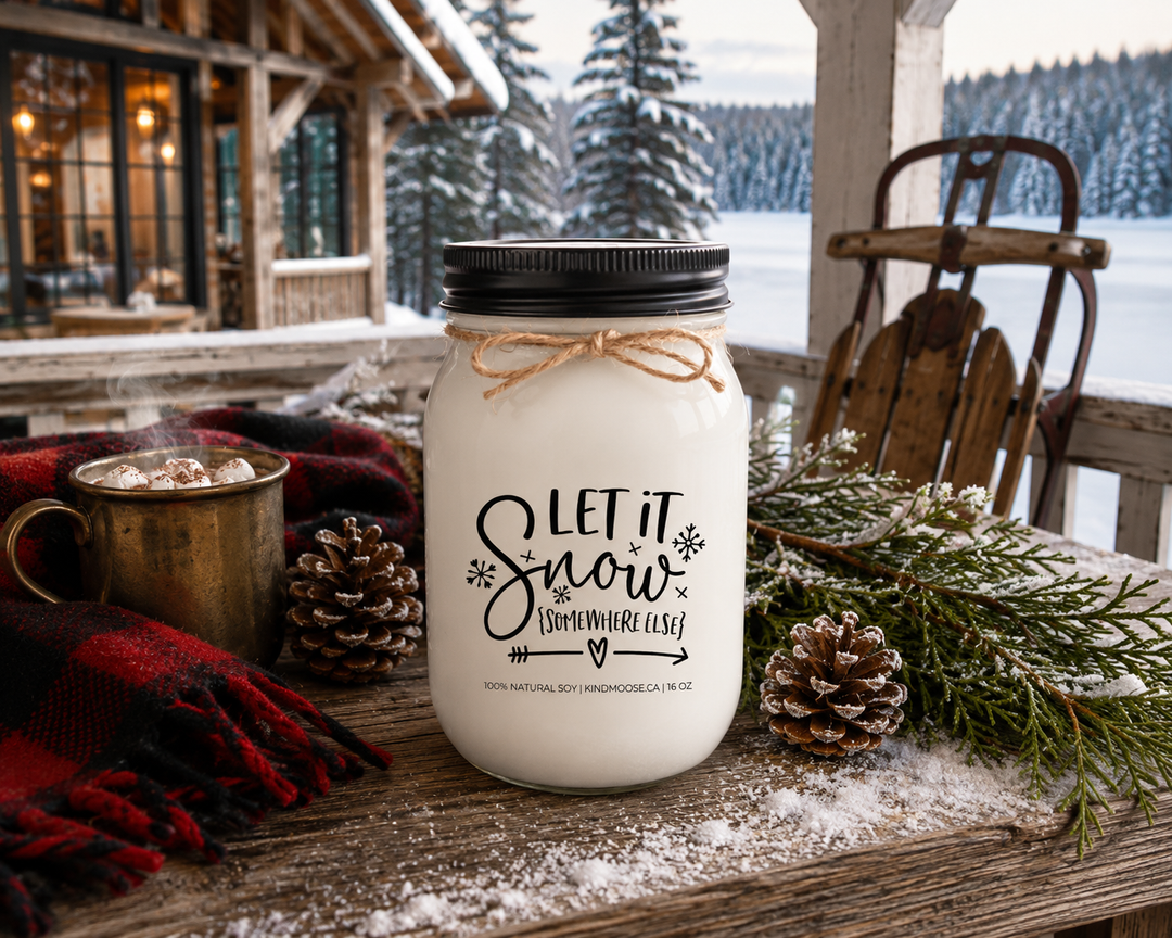 LET IT SNOW (SOMEWHERE ELSE) customizable KINDMOOSE soy candle with a black lid displayed on a reclaimed white oak table on a luxury Canadian lakeside cottage porch overlooking a frozen lake. Fresh cedar branches, pinecones, a steaming antique brass mug, buffalo plaid scarf, and vintage wooden sled create a warm, cozy winter retreat after a fresh snowfall.