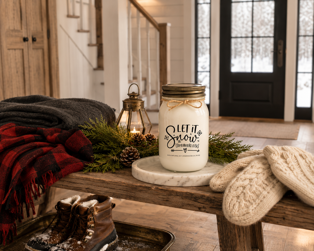 LET IT SNOW (SOMEWHERE ELSE) customizable KINDMOOSE soy candle with a distressed bronze lid resting on a reclaimed white oak entry bench inside a luxury Canadian farmhouse foyer. Snow-dusted boots, knit mittens, buffalo plaid scarf, cedar branches, pinecones, and antique brass accents tell the story of returning inside after a snowy winter day.