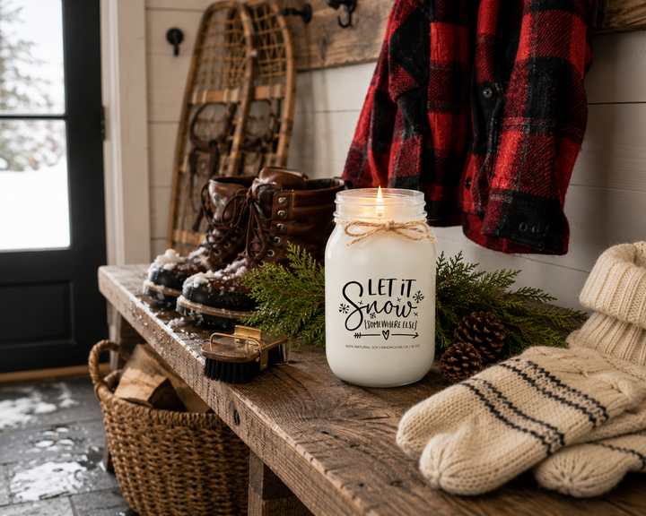 LET IT SNOW (SOMEWHERE ELSE) customizable KINDMOOSE soy candle burning without a lid on a reclaimed white oak mudroom bench inside a luxury Canadian lakeside cottage. Snow-covered boots, drying mittens, buffalo plaid jacket, vintage snowshoes, cedar branches, pinecones, and warm natural lighting celebrate the comfort of escaping the winter cold.