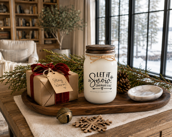 LET IT SNOW (SOMEWHERE ELSE) customizable KINDMOOSE soy candle with a distressed bronze lid displayed as the centerpiece of an elegant winter gift arrangement on a reclaimed white oak library console. Wrapped kraft gift with red velvet ribbon, cedar branches, pinecones, antique brass sleigh bell, carved snowflake ornament, and luxury Canadian cottage styling create the perfect cozy holiday gift scene.