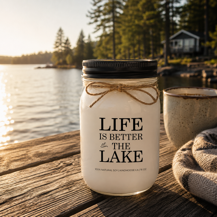 LIFE IS BETTER AT THE LAKE soy candle with black lid displayed on a weathered wooden dock overlooking a peaceful Canadian lake surrounded by pine trees and cottage scenery.