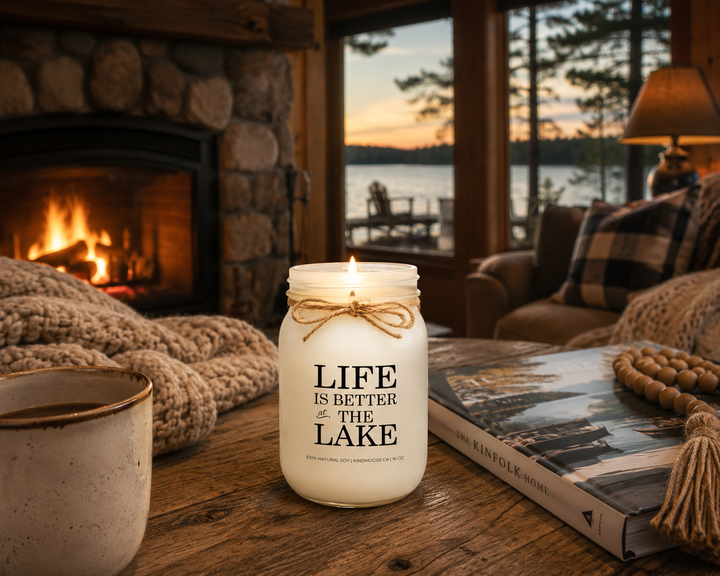 LIFE IS BETTER AT THE LAKE candle burning beside a stone fireplace inside a cozy lakeside cottage with rustic wood furniture, soft blankets, and lake views through large windows.