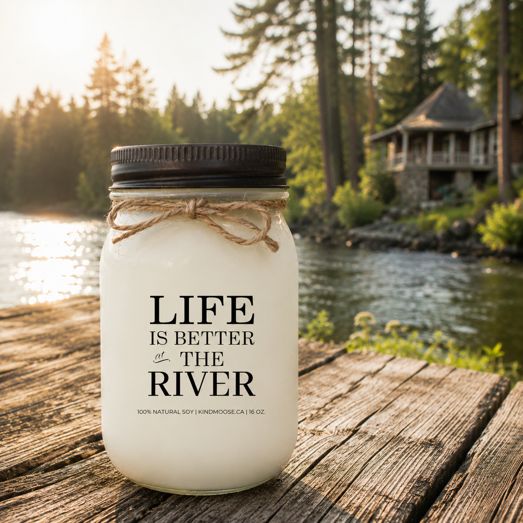 Life Is Better At The River 16 oz soy candle on a rustic wooden deck overlooking a peaceful river with morning sunlight and cottage scenery
