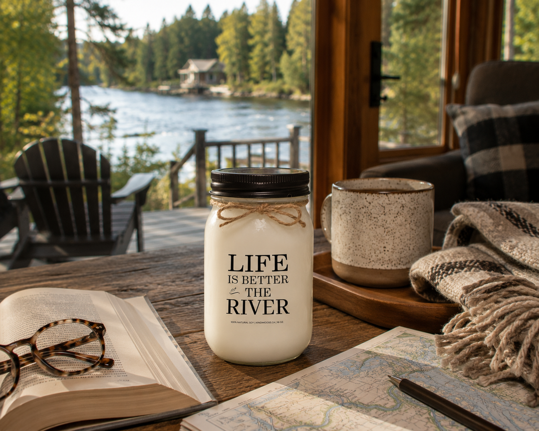 Life Is Better At The River candle with open novel, coffee mug and blanket beside a quiet river cottage setting