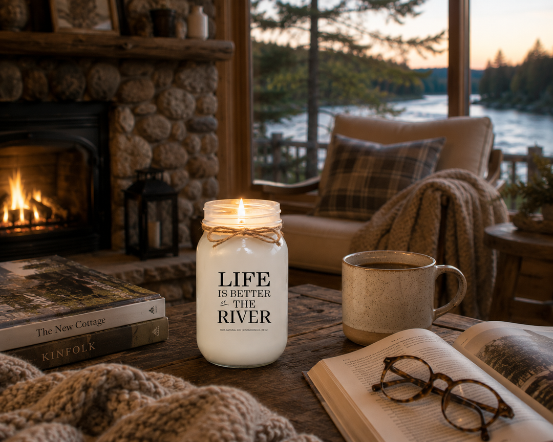 Life Is Better At The River soy candle burning inside a cozy riverside cottage with fireplace, warm lighting and peaceful river views