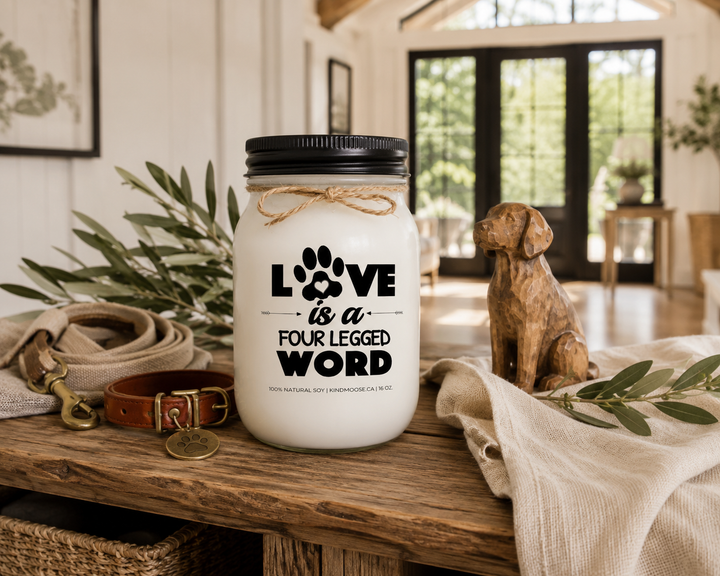 LOVE IS A FOUR LEGGED WORD customizable scented soy candle by KINDMOOSE Candle Co. displayed on a reclaimed white oak entryway console with a black lid, natural twine bow, leather dog collar, antique brass pet tag, carved wooden dog figurine, olive branches, and warm afternoon sunlight in a luxury Canadian modern farmhouse.