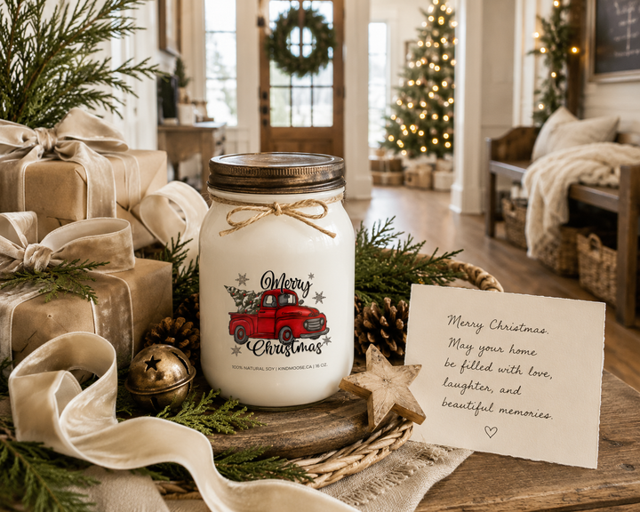 MERRY CHRISTMAS personalized farmhouse candle gift displayed on a rustic entry console with wrapped presents, evergreen branches, pinecones, a wooden star ornament, brass sleigh bell, ivory velvet ribbon, and a handwritten Christmas card. A frosted white KINDMOOSE mason jar candle with a distressed bronze lid sits as the centerpiece in a bright farmhouse foyer with a Christmas tree, wreath, and warm holiday décor, creating an elegant Christmas gifting scene.