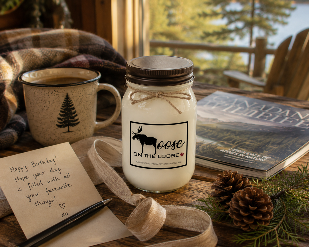 LIFE IS BETTER AT THE RIVER candle styled as a thoughtful gift with a handwritten card, linen ribbon, coffee mug, river lifestyle magazine, greenery, and warm golden sunlight on a rustic wood table.