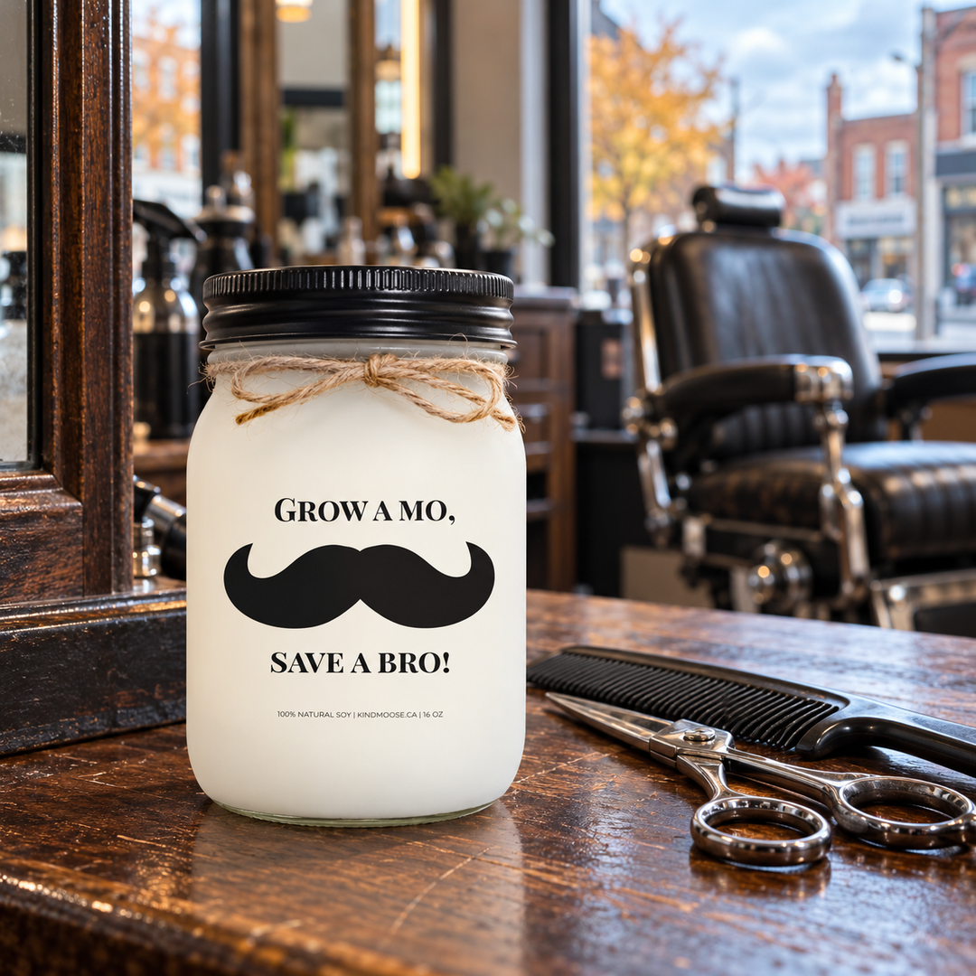 KINDMOOSE Grow a Mo, Save a Bro! 16 oz Movember soy candle with black lid and moustache label on a wooden workstation in a neighbourhood barbershop.