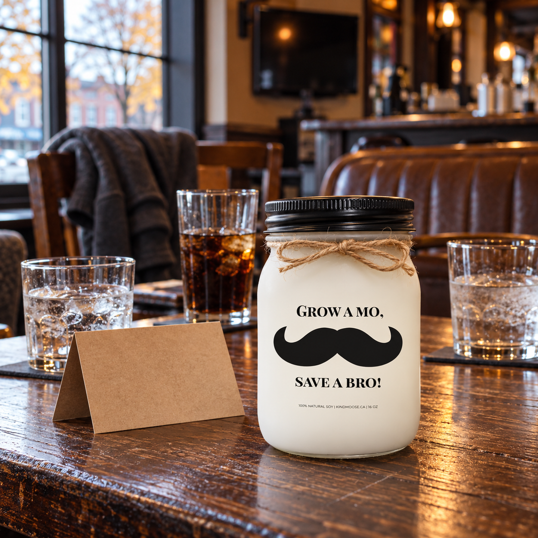 KINDMOOSE Grow a Mo, Save a Bro! Movember candle on a wooden pub table with three drinks, a greeting card and chairs suggesting a gathering of longtime friends.