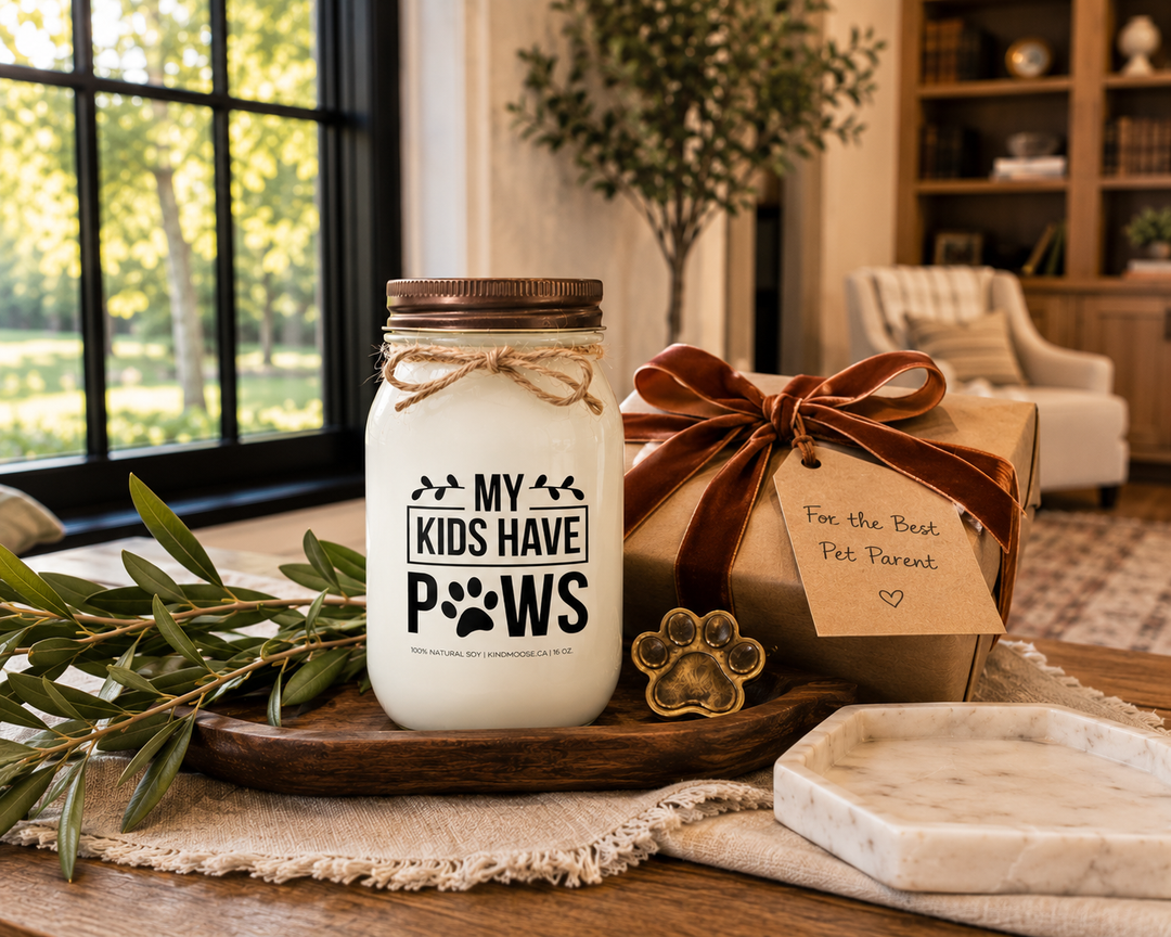 My Kids Have Paws customizable 16 oz soy candle with a distressed bronze lid displayed beside a beautifully wrapped kraft paper gift tied with a cognac velvet ribbon and gift tag reading "For the Best Pet Parent." A brass paw ornament, walnut tray, marble tray, olive branches, and warm afternoon sunlight create an elegant modern farmhouse pet-lover gift scene.