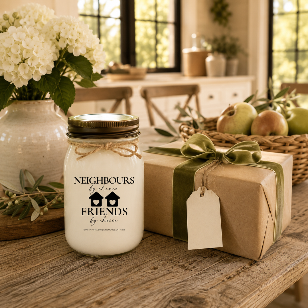 Neighbours by Chance, Friends by Choice customizable 16 oz soy candle with a distressed bronze lid displayed beside a beautifully wrapped housewarming gift with a sage velvet ribbon and blank gift tag. White hydrangeas, fresh pears, apples, and olive branches create an elegant modern farmhouse gift scene.