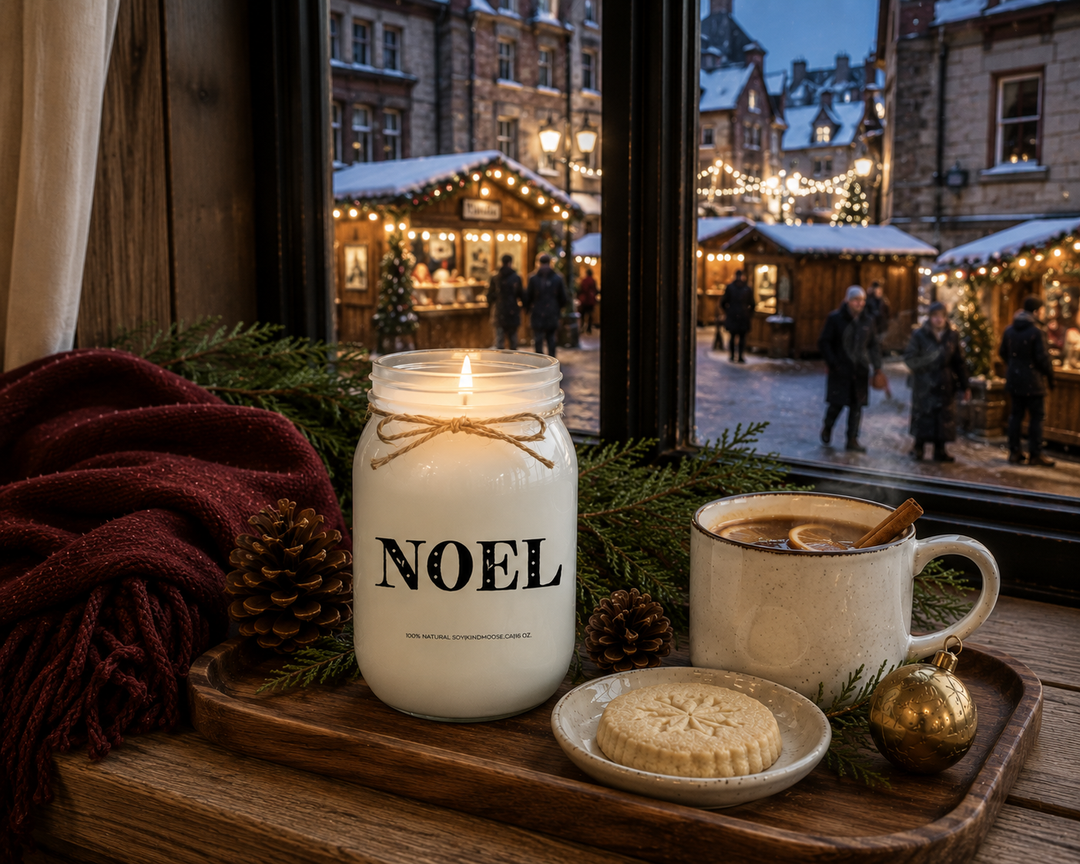 Lit NOEL customizable Christmas candle on a reclaimed white oak window seat overlooking a softly glowing European Christmas market at blue hour, styled with a burgundy cashmere throw, mulled cider, shortbread cookie, cedar sprigs, and pinecones for a peaceful and magical holiday evening.