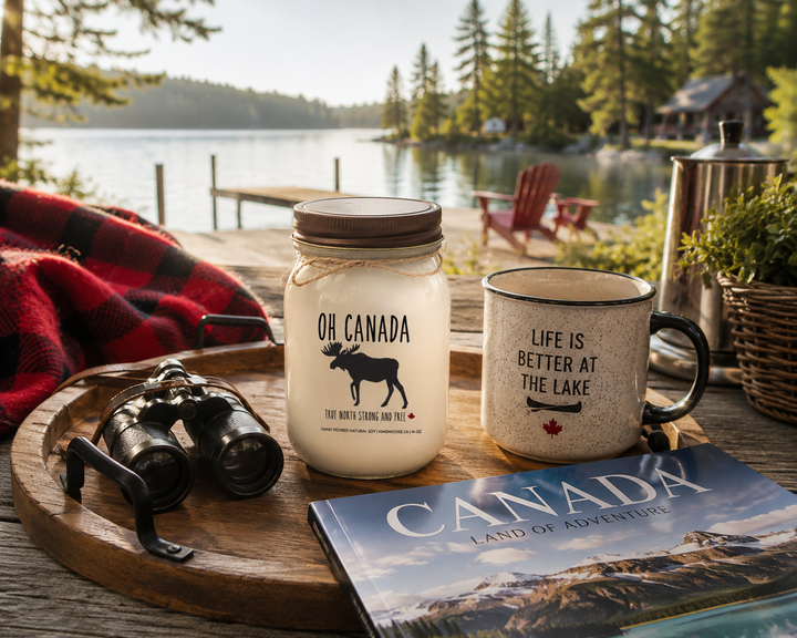 OH CANADA soy candle with distressed bronze lid styled on a cottage deck beside a coffee mug, Canadian travel magazine, plaid blanket, and binoculars overlooking a lakeside view.