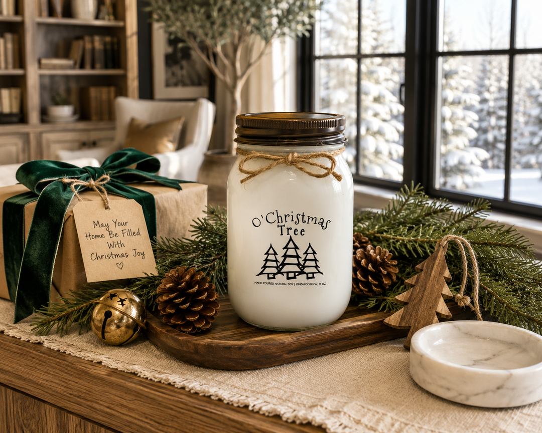 O' Christmas Tree customizable 16 oz soy candle with distressed bronze lid displayed as an elegant Christmas gift on a reclaimed white oak library console, styled with a beautifully wrapped kraft paper present, deep forest green velvet ribbon, Fraser fir branches, pinecones, antique brass sleigh bell, carved wooden Christmas tree ornament, and softly falling snow outside large black-framed windows.