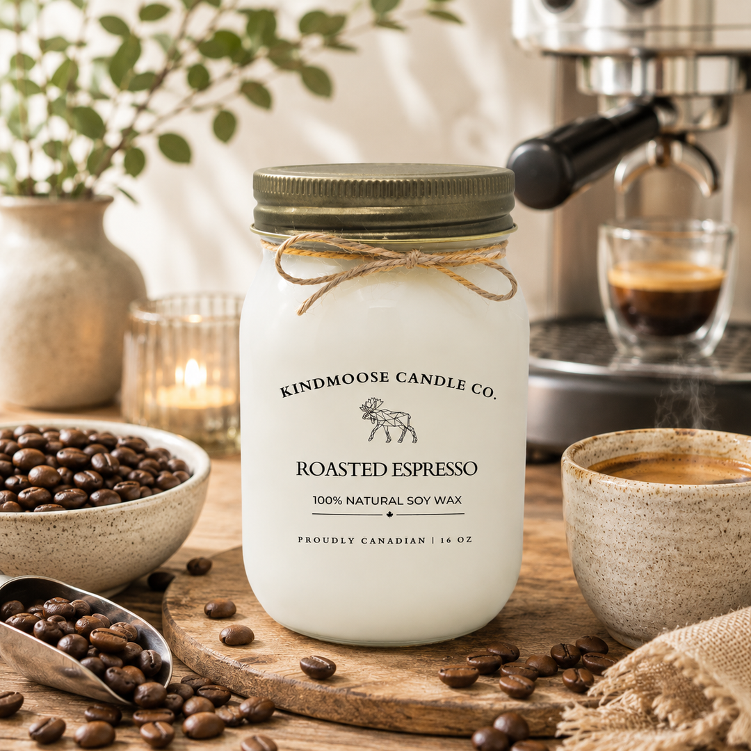 KINDMOOSE Candle Co. Roasted Espresso 16 oz soy candle displayed on a rustic wooden board beside freshly roasted coffee beans and a steaming espresso cup. Warm morning sunlight fills the cozy coffeehouse-inspired setting, while a softly blurred espresso machine and natural greenery create an inviting atmosphere. The frosted white candle jar with twine detail stands as the focal point, evoking the rich aroma and comforting experience of freshly brewed espresso.