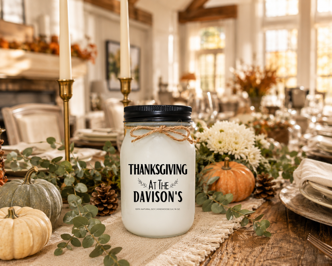 Personalized "THANKSGIVING AT THE..." KINDMOOSE 16 oz soy candle with a black lid displayed as the centerpiece of a beautifully decorated farmhouse Thanksgiving table. Surrounded by heirloom pumpkins, eucalyptus garland, white chrysanthemums, pinecones, neutral dinnerware, and warm autumn décor, the candle celebrates family traditions, gratitude, and gathering around the table.