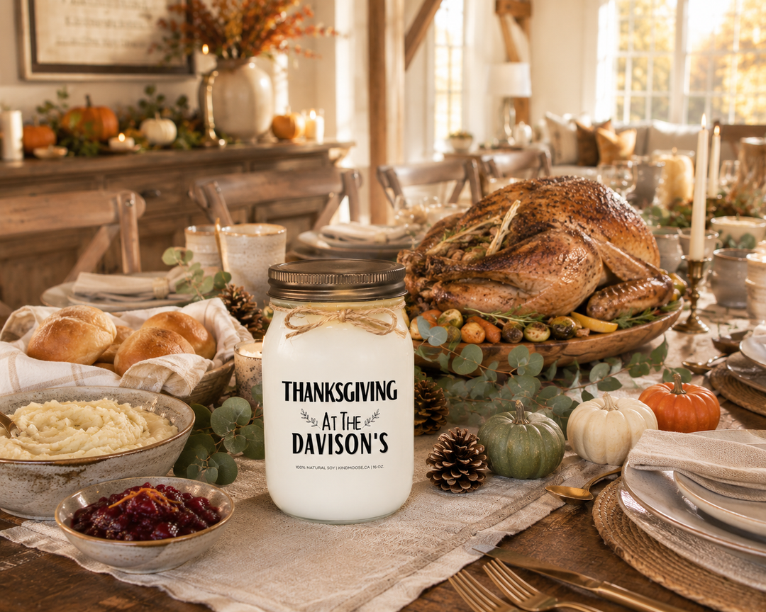 Personalized "THANKSGIVING AT THE..." KINDMOOSE 16 oz soy candle with a distressed bronze lid displayed on a rustic farmhouse Thanksgiving table prepared for family dinner. Roasted turkey, mashed potatoes, dinner rolls, cranberry sauce, seasonal vegetables, heirloom pumpkins, and elegant harvest décor create a warm, welcoming atmosphere before guests arrive.