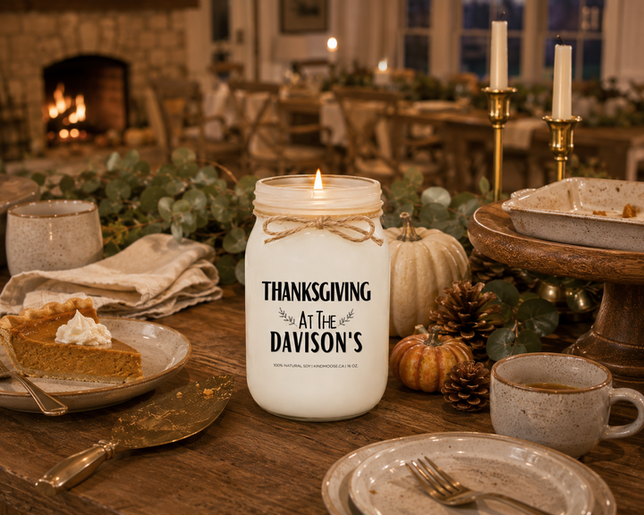 Personalized "THANKSGIVING AT THE..." KINDMOOSE 16 oz soy candle burning without a lid on a rustic farmhouse sideboard after Thanksgiving dinner. A slice of pumpkin pie, coffee cups, folded linen napkins, heirloom pumpkins, pinecones, and soft evening lighting create a peaceful scene celebrating family memories and gratitude after the holiday meal.