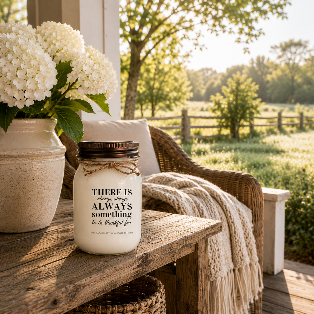 KINDMOOSE "There Is Always Something to Be Thankful For" 16 oz soy candle on a peaceful farmhouse porch overlooking the countryside with hydrangeas, wicker seating, and a cozy knit blanket.