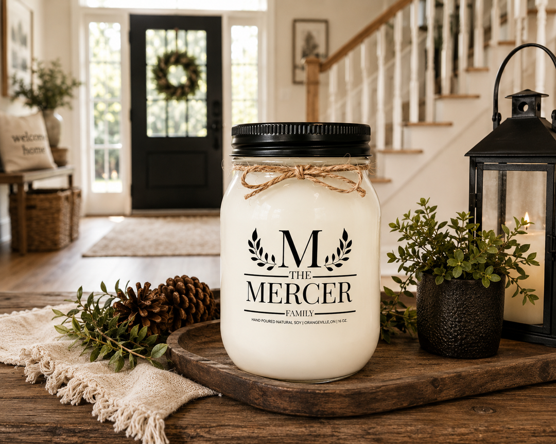 Customized Family Soy Candle displayed on a reclaimed wood entry console in a bright luxury farmhouse foyer. Personalized family name label is fully readable on a frosted white mason jar with black lid and twine bow, surrounded by natural wood textures, linen accents, and elegant farmhouse décor, creating a warm welcome-home atmosphere.